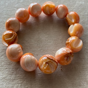 May include: A bracelet made of round, peach-colored beads with swirling patterns. The beads are made of a material that resembles mother-of-pearl.