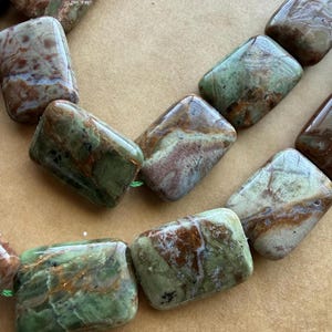 May include: A strand of polished, rectangular beads in shades of green, brown, and cream. The beads are strung together with green thread. The natural stone beads have a glossy finish and varying patterns, suggesting a unique, handcrafted jewelry component.