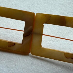 Op de afbeelding: Twee rechthoekige, amberkleurige gespen met een vierkante uitsparing in het midden. Een dun, oranje draadje loopt door het midden van elke gesp. De gespen hebben een gemarmerd uiterlijk.