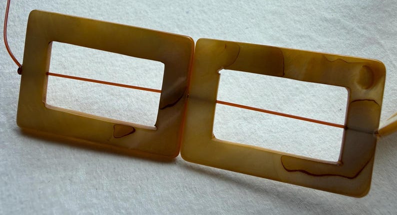 Op de afbeelding: Twee rechthoekige, amberkleurige gespen met een vierkante uitsparing in het midden. Een dun, oranje draadje loopt door het midden van elke gesp. De gespen hebben een gemarmerd uiterlijk.