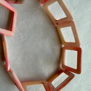 Op de afbeelding: Een ketting met vierkante, open kralen in roze en perziktinten. De kralen zijn aan elkaar geregen, waardoor een geometrisch patroon ontstaat. De ketting wordt getoond tegen een witte achtergrond.