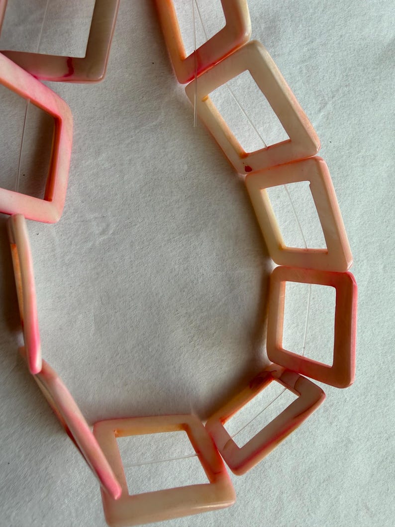 Op de afbeelding: Een ketting met vierkante, open kralen in roze en perziktinten. De kralen zijn aan elkaar geregen, waardoor een geometrisch patroon ontstaat. De ketting wordt getoond tegen een witte achtergrond.