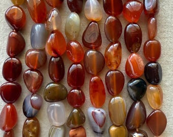 Op de afbeelding: Een verzameling gepolijste, getrommelde edelsteenkralen gerangschikt in verticale strengen. De stenen vertonen een verscheidenheid aan warme kleuren, waaronder tinten diep rood, gebrand oranje, amber, bruin en doorschijnend wit met natuurlijke banden.