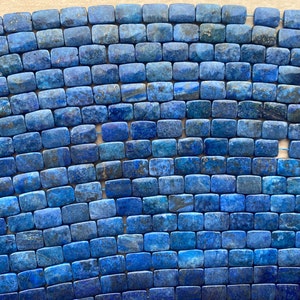 May include: A close-up of a strand of blue lapis lazuli beads. The beads are rectangular and have a smooth, polished finish. The lapis lazuli is a deep blue color with flecks of gold pyrite.