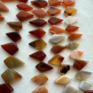 May include: A collection of polished, geometric gemstones in shades of orange, brown, and cream. The stones are diamond-shaped with a flat top and bottom, and are arranged on a white surface.