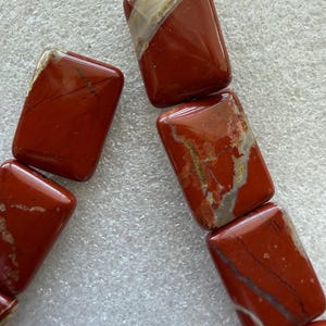 May include: Close-up of a strand of rectangular red jasper beads. The beads have a polished finish with natural veining in shades of white, tan, and gray. The beads are strung together, suggesting they are for jewelry making.