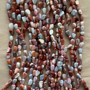May include: Strands of polished gemstone beads in shades of red, white, gray, and brown. The beads are oval and irregular in shape, strung together, and suitable for jewelry making or crafting projects.