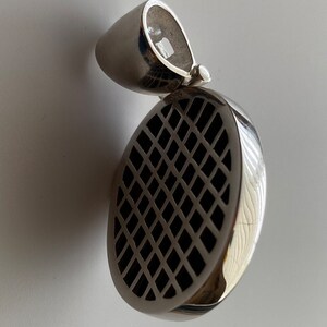 May include: Silver pendant with a round, black, grid-like design. The pendant has a loop at the top for attaching to a chain.
