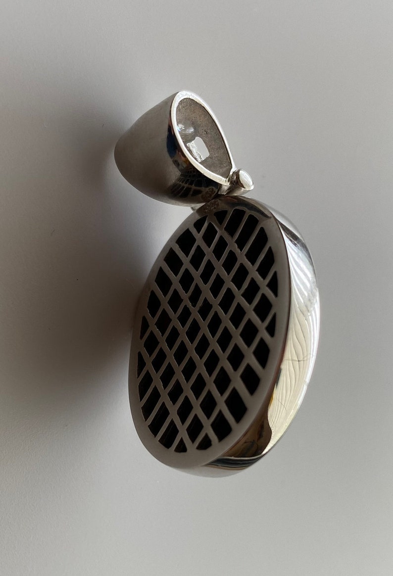 May include: Silver pendant with a round, black, grid-like design. The pendant has a loop at the top for attaching to a chain.