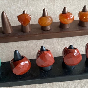 May include: A collection of ten faceted gemstone rings displayed on two rectangular stands. The top stand holds four orange carnelian rings, while the bottom stand features one green aventurine ring and four deep red jasper rings on black cones.