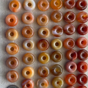 May include: A collection of 50 carnelian donut beads, each with a hole in the center. The beads are a mix of orange, red, and brown colors with white streaks.
