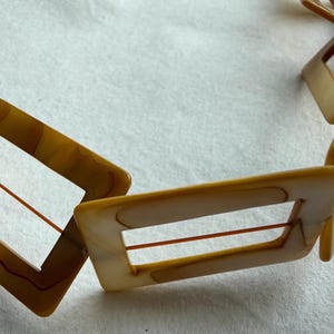 Op de afbeelding: Een vintage ketting met rechthoekige, open-frame schakels in tinten bruin en cr&egrave;me. De schakels zijn verbonden door dunne, roodbruine koorden. De ketting ligt op een witte ondergrond, wat het geometrische ontwerp en het warme kleurenpalet benadrukt.