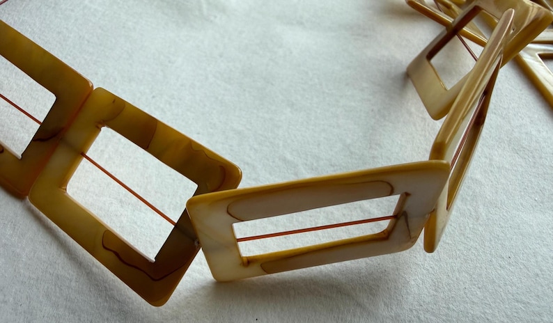 Op de afbeelding: Een vintage ketting met rechthoekige, open-frame schakels in tinten bruin en cr&egrave;me. De schakels zijn verbonden door dunne, roodbruine koorden. De ketting ligt op een witte ondergrond, wat het geometrische ontwerp en het warme kleurenpalet benadrukt.