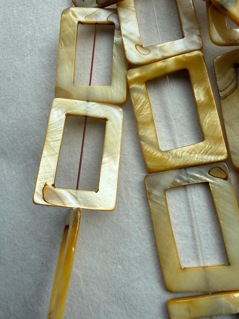Op de afbeelding: Close-up van verschillende rechthoekige, iriserende, cr&egrave;mekleurige gespen. Elke gesp heeft een rechthoekige opening en een koperkleurige metalen staaf. De gespen zijn diagonaal gerangschikt.