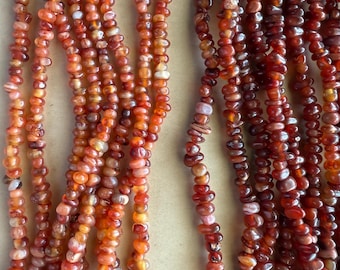 May include: Close-up of multiple strands of orange and reddish-brown gemstone beads. The beads are small, irregular in shape, and strung together. The color gradient ranges from light orange to deep reddish-brown.