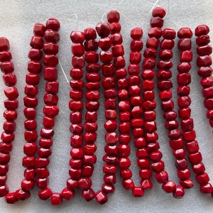 May include: Strands of vibrant red coral beads. The beads are a mix of shapes, including square and rounded, strung together with white string. The coral has a polished, glossy finish, reflecting light.
