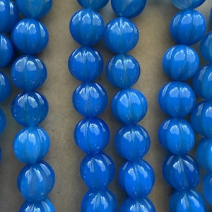 May include: Close-up of strands of bright blue, round beads. Each bead has a ribbed texture, giving them a unique, segmented appearance. The beads are strung together, creating a pattern of vertical lines. The color is a vibrant, solid blue.