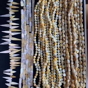 May include: Assortment of yellow and white shell beads in various shapes and sizes. The beads are arranged in rows on a black background. The beads are perfect for jewelry making.