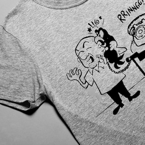 Puede incluir: Una camiseta gris con una ilustración de dibujos animados en blanco y negro de una persona que se sobresalta por un teléfono que suena. El texto "RRRINNGG!" está encima del teléfono.