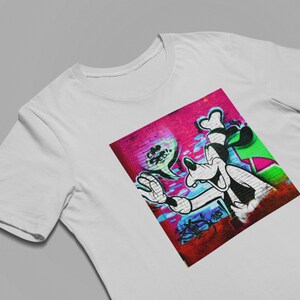 May include: A white t-shirt with a colorful graphic print of a cartoon dog with a goofy grin. The dog is painted on a brick wall with pink, blue, green, and white paint.