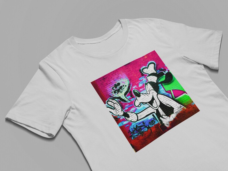 May include: A white t-shirt with a colorful graphic print of a cartoon dog with a goofy grin. The dog is painted on a brick wall with pink, blue, green, and white paint.