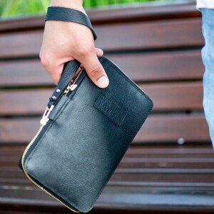 Könnte beinhalten: Eine schwarze Leder-Clutch mit Reißverschluss aus Gold und einem kleinen geprägten Logo.