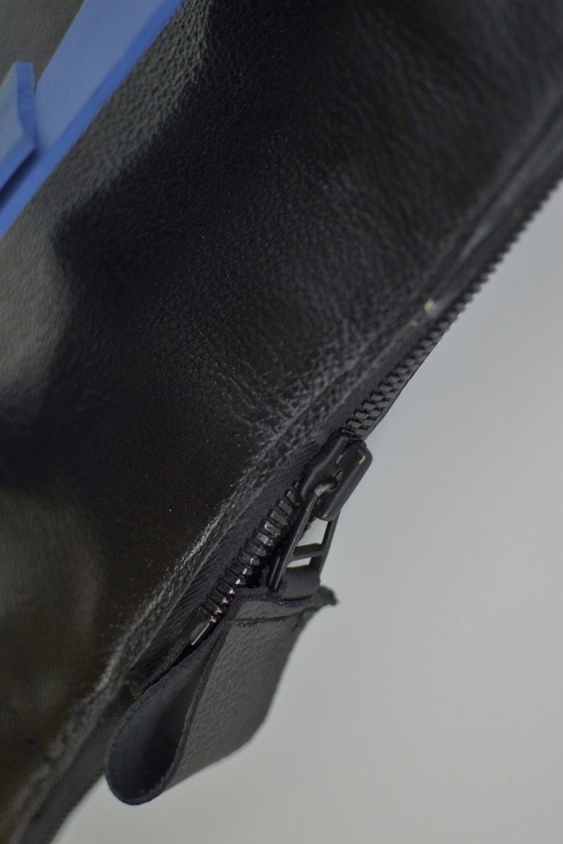 Puede incluir: Primer plano de una cremallera de cuero negro con una leng&uuml;eta azul. La cremallera est&aacute; en una bolsa de cuero negro.