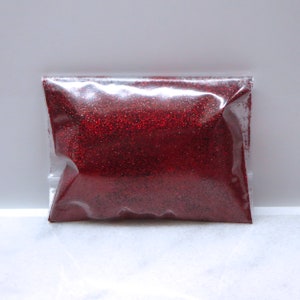 May include: A clear plastic bag filled with vibrant red glitter. The glitter is finely ground and reflects light, creating a sparkling effect. The bag is sealed and sits on a white surface, showcasing the glitter's color and texture.