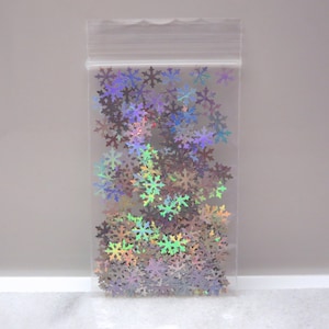 May include: A clear plastic bag filled with iridescent snowflake-shaped confetti. The confetti reflects a range of colors, including silver, purple, and green. The bag is sealed at the top, and the snowflakes are scattered throughout.