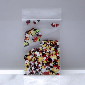 May include: A clear plastic bag filled with colorful Mickey Mouse head shaped glitter. The glitter is red, yellow, black, and white.