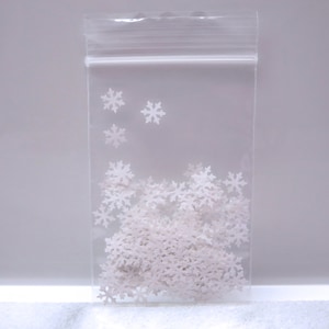 May include: A clear plastic bag filled with white snowflake-shaped confetti. The bag is sealed at the top and contains a large quantity of the snowflake decorations. The snowflakes are scattered throughout the bag.