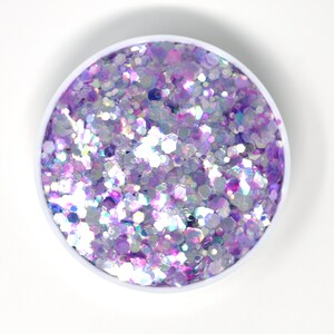 May include: A close-up of a small container filled with iridescent purple and white glitter. The glitter is a mix of different sizes and shapes, creating a sparkly and festive look.
