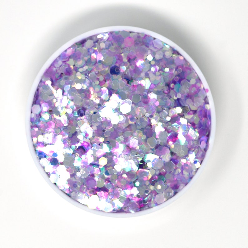 May include: A close-up of a small container filled with iridescent purple and white glitter. The glitter is a mix of different sizes and shapes, creating a sparkly and festive look.