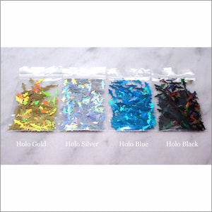 May include: Four small bags of holographic shark shaped glitter. The bags are labeled "Holo Gold", "Holo Silver", "Holo Blue", and "Holo Black".