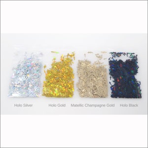 May include: Four small plastic bags filled with cross-shaped glitter in various colors. The colors include holo silver, holo gold, metallic champagne gold, and holo black. The names of the colors are printed below each bag.
