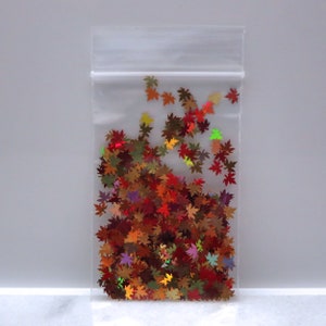 May include: A clear plastic bag filled with colorful, maple leaf-shaped confetti. The confetti features shades of red, orange, yellow, and green, creating a fall-themed aesthetic. The bag is sealed at the top.