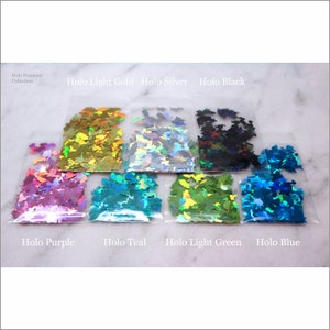 May include: Assortment of holographic butterfly shaped glitter in various colors, including purple, teal, light green, blue, light gold, silver, and black. The glitter is packaged in clear plastic bags with the color name printed below each bag.