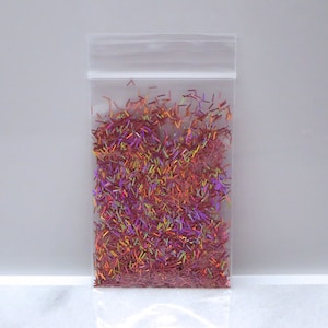 May include: A clear plastic bag filled with iridescent glitter. The glitter is composed of small, colorful, rod-shaped pieces in shades of pink, purple, orange, and green. The bag is sealed at the top.
