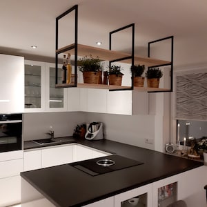 May include: A white kitchen with a black countertop and a black hob. There are black metal shelves hanging from the ceiling with wooden shelves and potted plants.