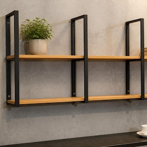 Op de afbeelding: Een wandplank met twee niveaus, houten planken en zwarte metalen frames. Een kleine plant in een pot en boeken worden op de planken tentoongesteld. De plank is aan een grijze muur bevestigd.