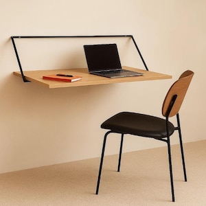 Peut inclure: Un bureau minimaliste mural en bois clair, avec un ordinateur portable et un carnet. Une chaise en bois avec une assise noire et des pieds en métal noir est placée à côté. Le bureau est soutenu par une structure en métal noir.