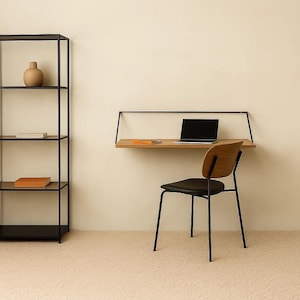 Pode incluir: Uma configuração moderna de escritório em casa com uma secretária flutuante de madeira com estrutura de metal preta, um laptop e uma cadeira. Uma estante de metal preta com objetos decorativos está à esquerda. A cadeira tem assento preto e encosto de madeira.