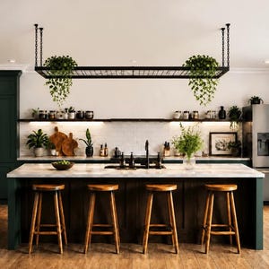 Op de afbeelding: Een keuken met een groot kookeiland met een wit aanrechtblad en vier houten krukken. Boven het eiland hangt een zwart metalen rek met hangende groene planten. Er zijn donkergroene kasten, een roestvrijstalen koelkast en open planken aanwezig.