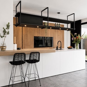 Könnte beinhalten: Eine moderne Küche mit einer weißen Kücheninsel, einer hellen Holzplatte und zwei schwarzen geflochtenen Barhockern. Über der Insel befindet sich ein schwarzes, von der Decke hängendes Metallregal mit integrierter Beleuchtung vor einer Holzwand.