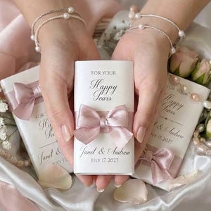 Könnte beinhalten: Weiße rechteckige Taschentuchpackungen mit dem Text FOR YOUR Happy Tears und Janet & Andrew JULY 17 2027. Jede Packung ist mit einer altrosa Satin-Schleife verziert. Die Artikel sind mit hellrosa Rosen und Perlenketten arrangiert.