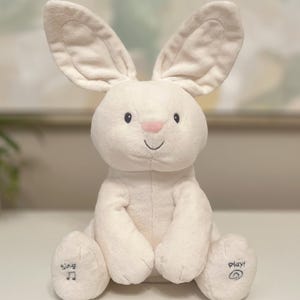 May include: A plush white bunny toy with long ears, a pink nose, and a smiling mouth. The bunny has the word "Sing" and a musical note on one foot, and "Play!" with a circular symbol on the other.