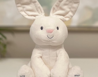 May include: A plush white bunny toy with long ears, a pink nose, and a smiling mouth. The bunny has the word "Sing" and a musical note on one foot, and "Play!" with a circular symbol on the other.