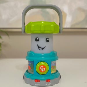 May include: A teal and green toy lantern with a smiling face and a gray handle. The lantern has a green top, a teal base, and a gray bottom. The lantern has the letters "fp" on the front. The lantern has buttons and illustrations of scenes.