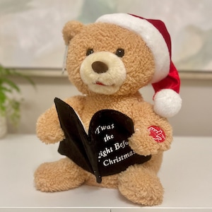 May include: A plush teddy bear wearing a Santa hat, holding a black book with the text "'Twas the night Before Christmas." The bear is light brown with a red button on its paw.