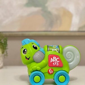 May include: A green and blue toy car shaped like a smiling chameleon. The toy has blue wheels, a pink panel with "ABC 123", and the "fp" logo. The chameleon has a blue crest and a gray textured area.